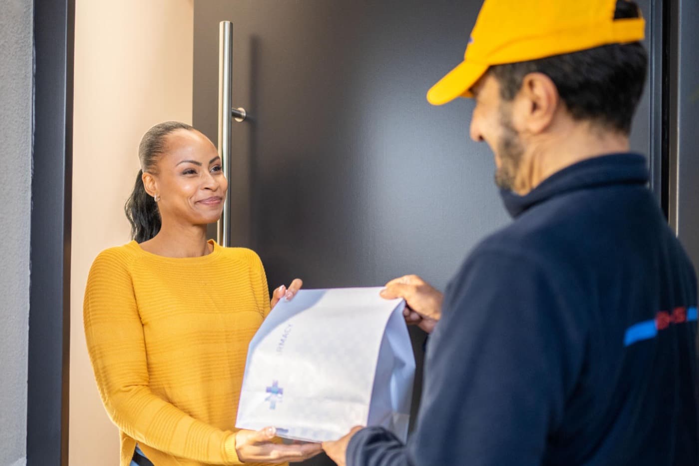 A smiling woman receiving a sealed pharmacy bag from a courier at her front door